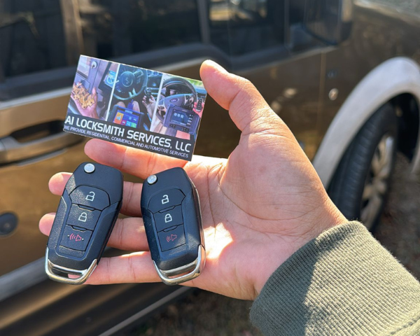 A hand holds an A1 Locksmith Services, LLC business card alongside two newly programmed automotive flip key fobs in front of a vehicle, highlighting professional mobile automotive locksmith services